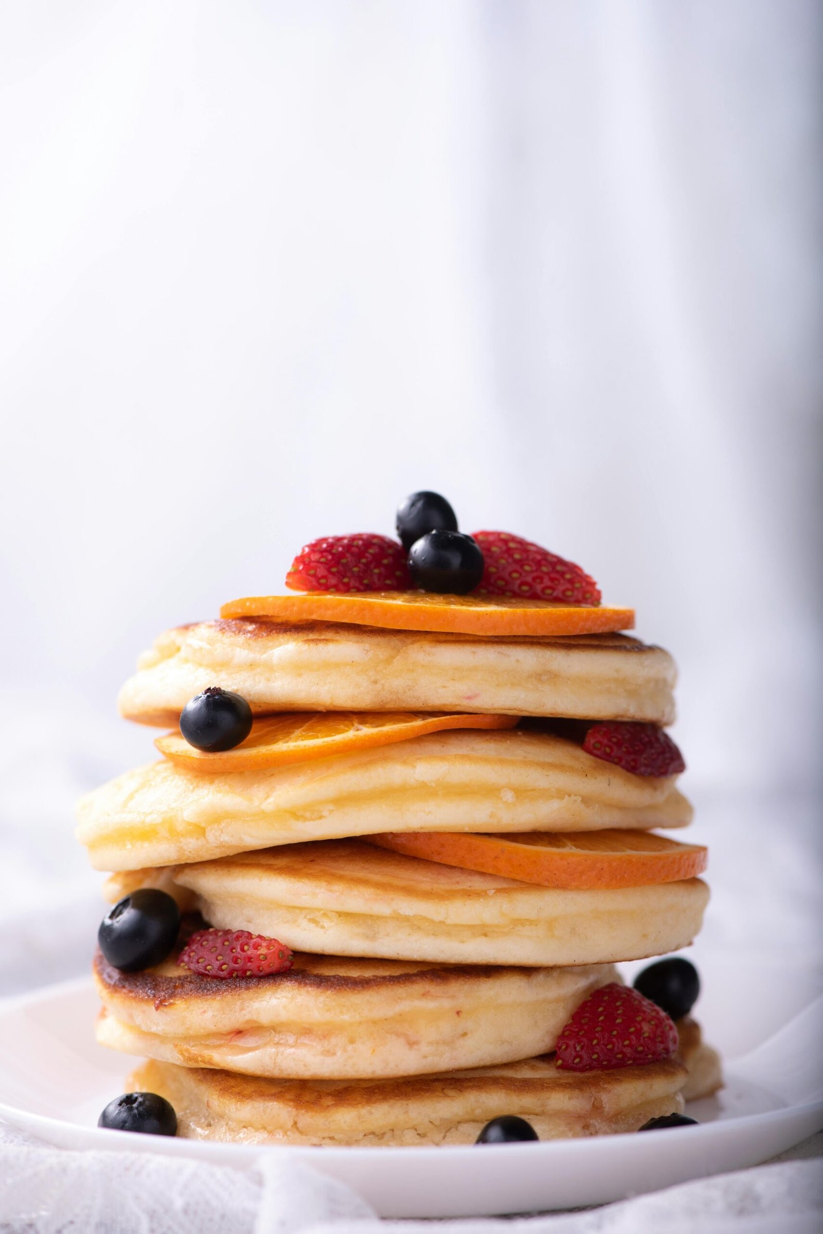 Tall stack of fluffy pancakes topped with blueberries, strawberries, and orange slices.